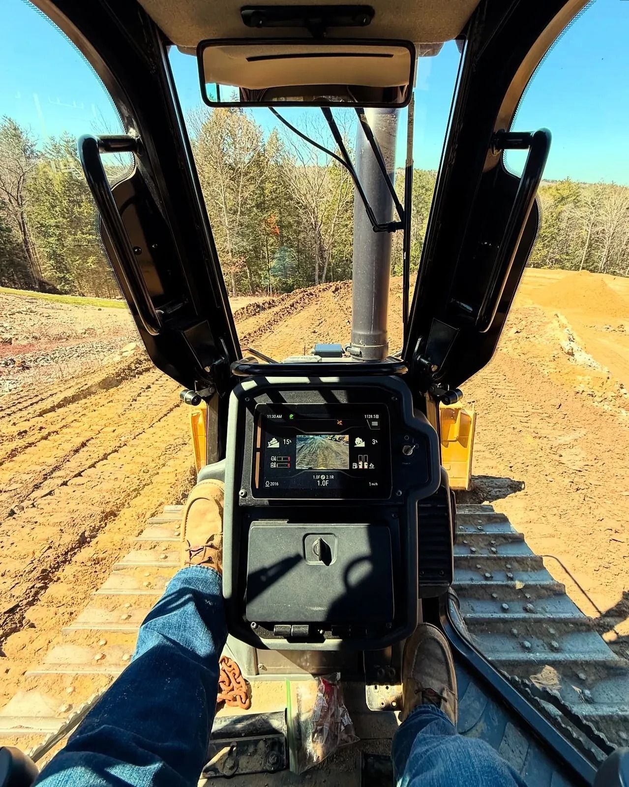 View from inside a bulldozer cab, looking out at a dirt track. A person's legs and feet are visible.