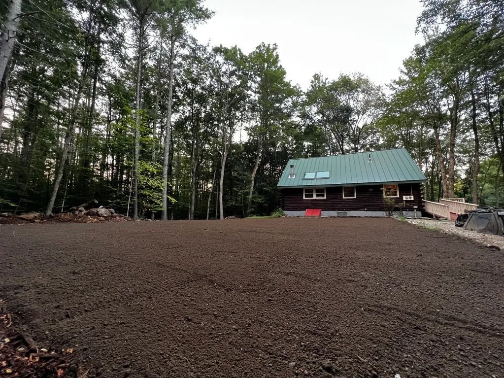 Log cabin with green roof amidst trees, in front of a recently tilled, dark brown yard.