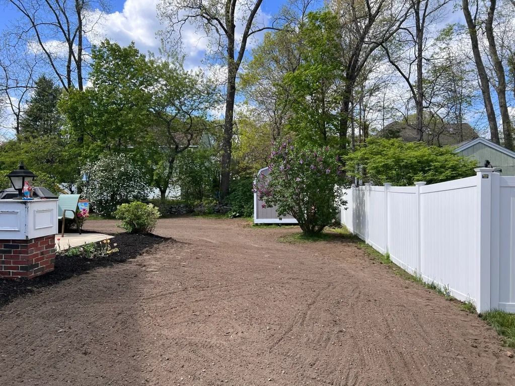 Gravel-covered yard with white fence and shrubs. Trees and cloudy sky in background.