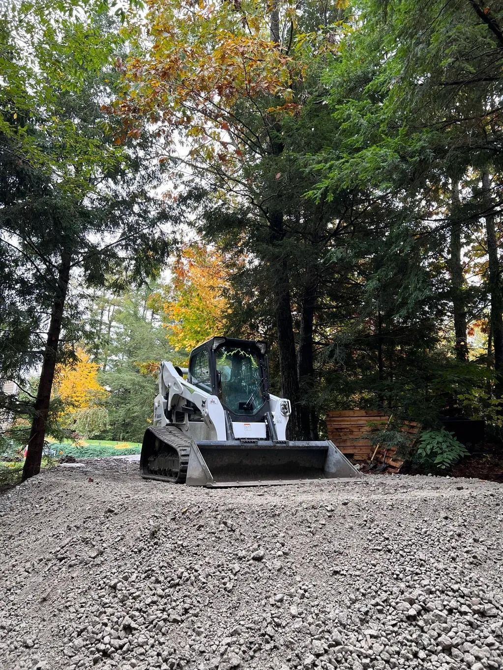 Bobcat skid-steer loader on a gravel pile in a wooded area, surrounded by green and yellow trees.