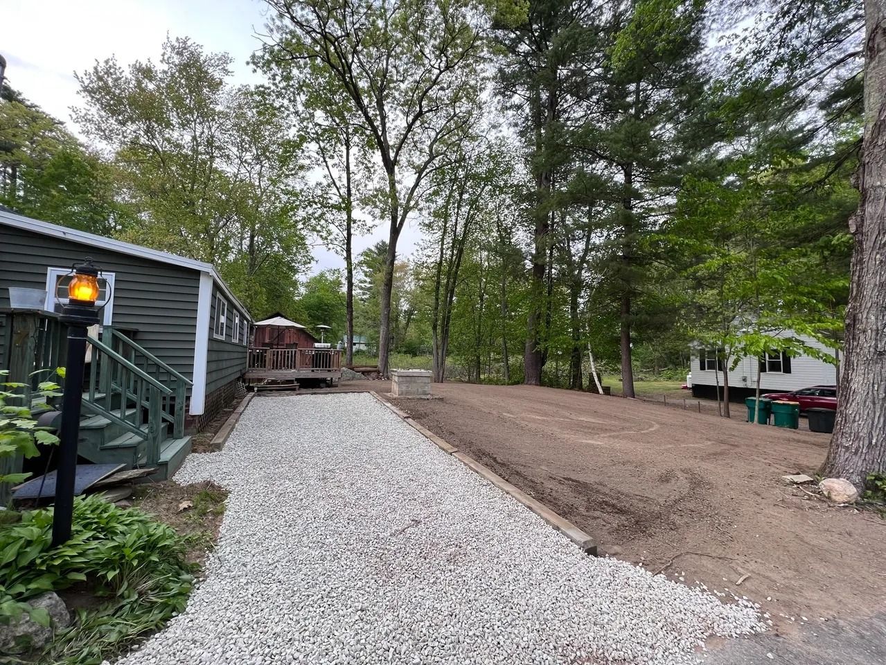 Gravel driveway leads to a wooden house with trees in the background, a light post on the left.