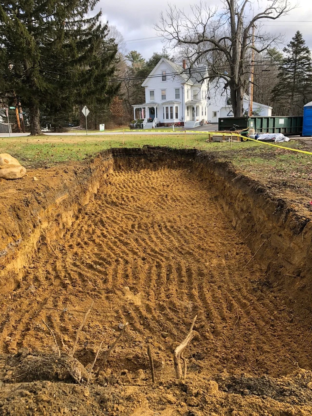 Large rectangular excavation in a grassy area with a white house in the background. Brown dirt and tire tracks visible.