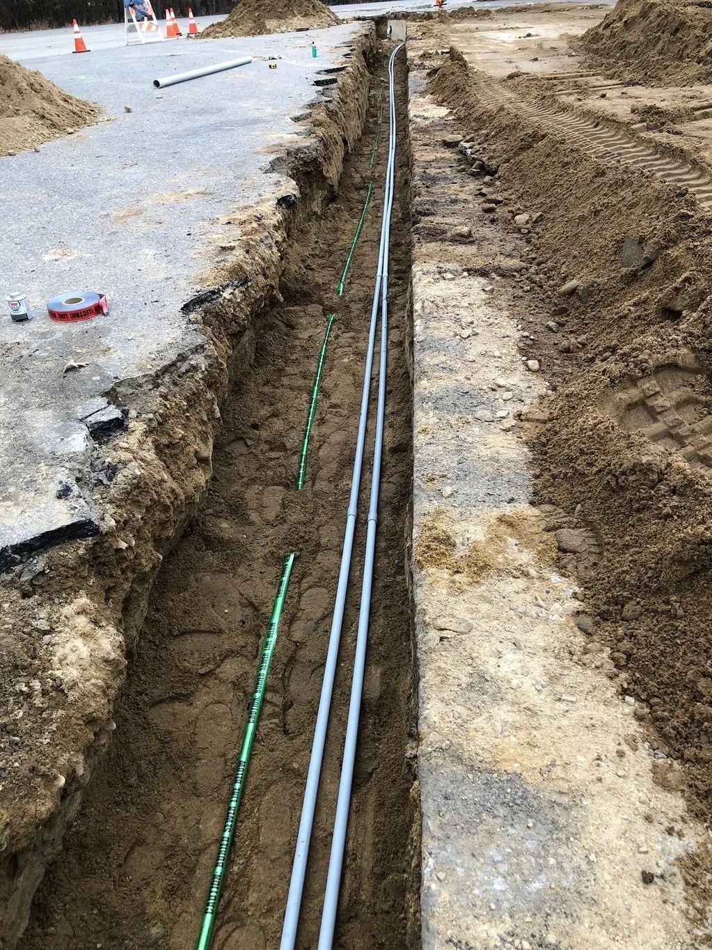 A long trench dug in the ground, holding gray and green pipes, beside a road.