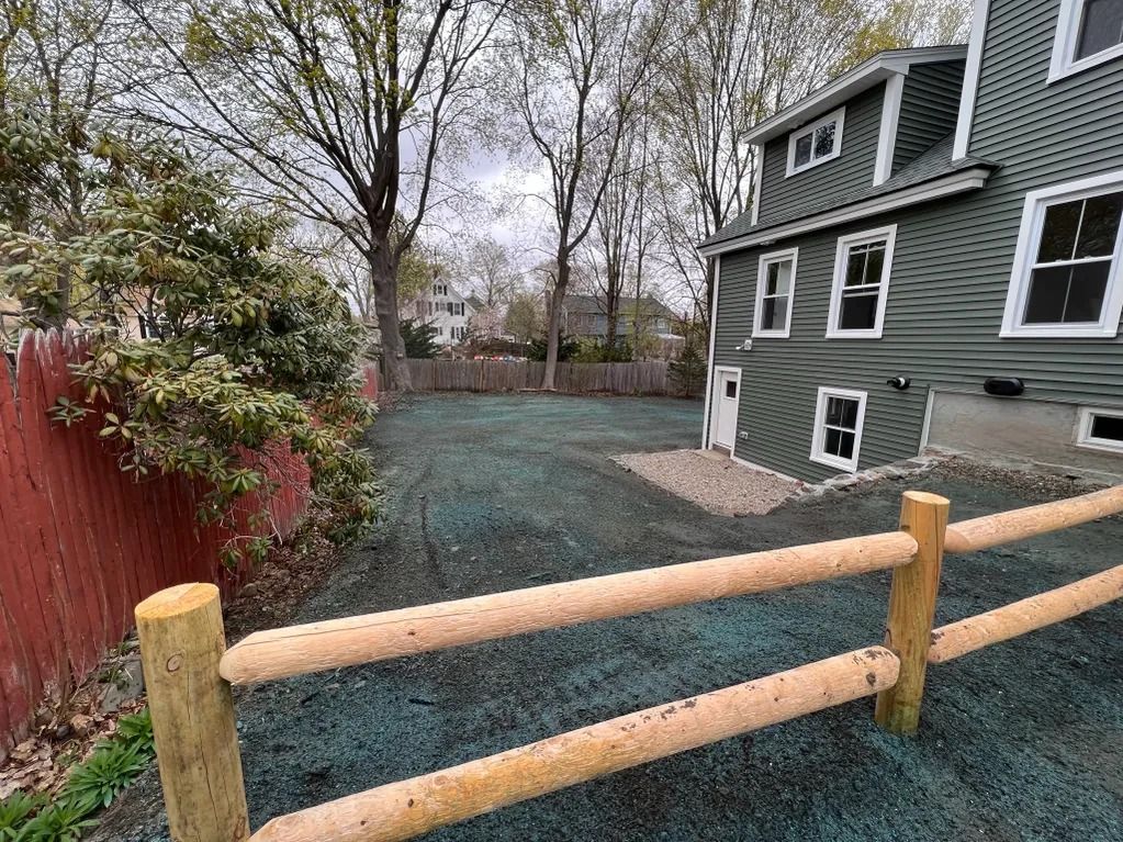 A backyard with a wooden fence, green lawn, and a two-story house with green siding and white-framed windows.