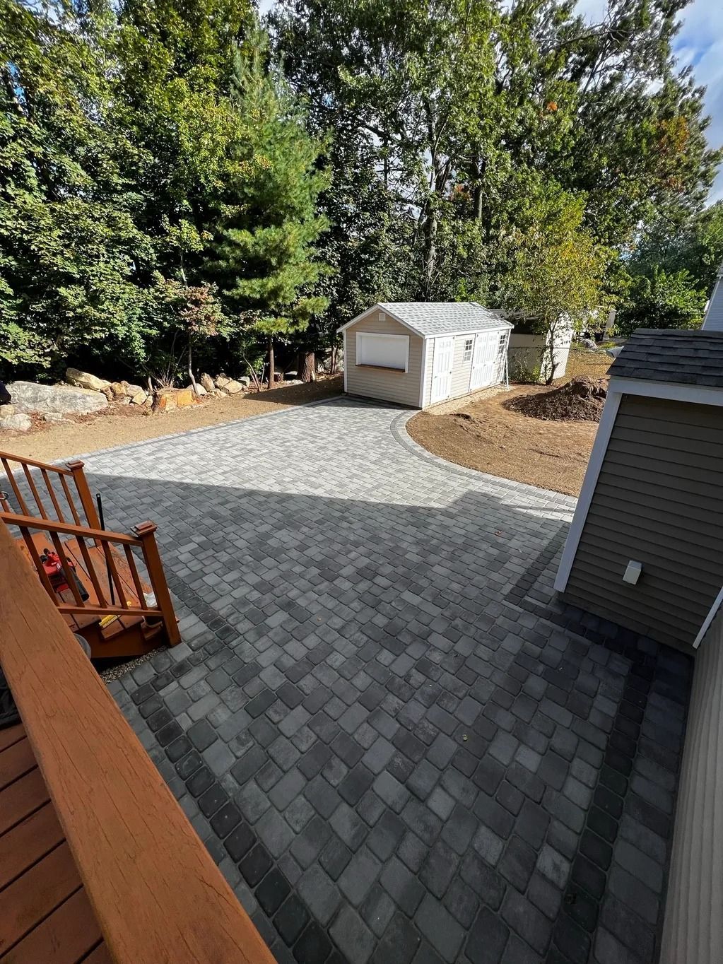 Patio with gray pavers, leading to a small, white shed. Wooden deck and trees in the background.