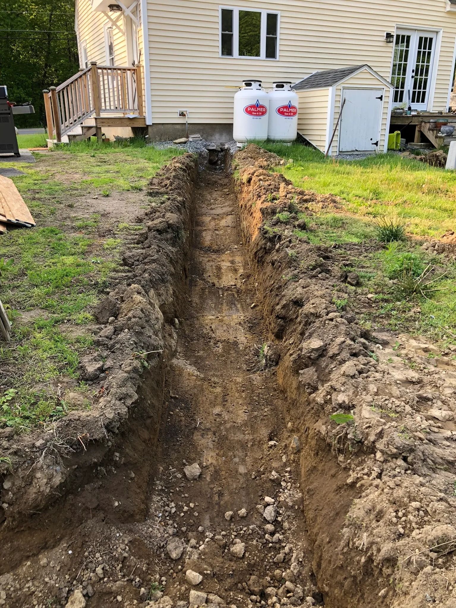 Trench dug in a grassy yard, leading toward a house. Two white propane tanks visible behind the trench.
