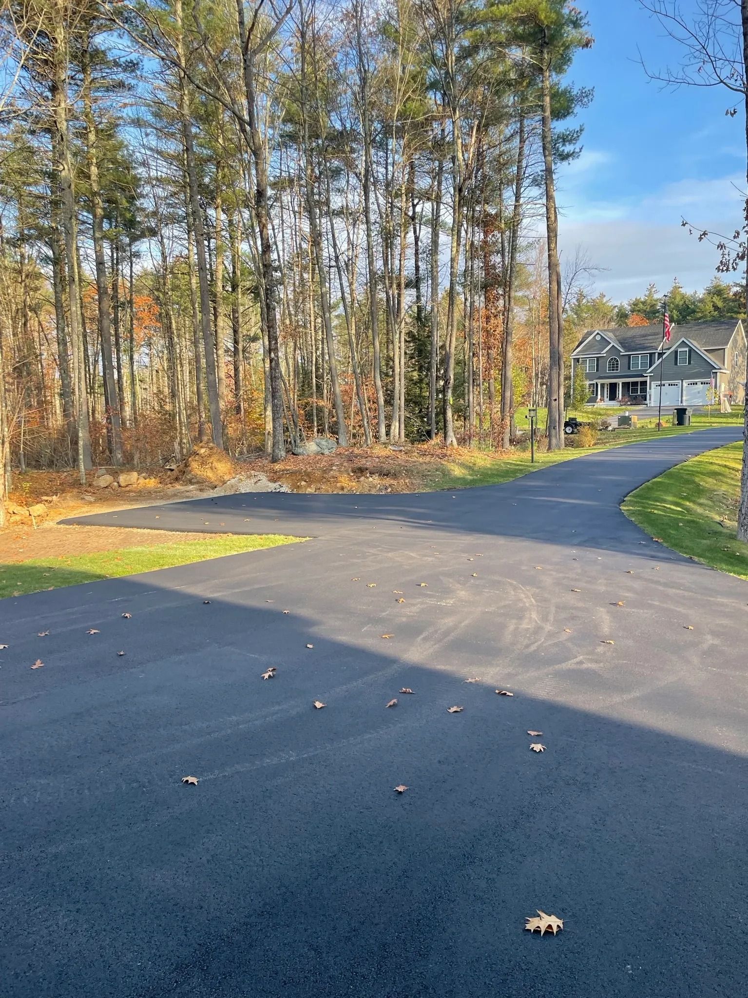 Paved road curves towards a house, with trees on the left and a blue sky in the background.