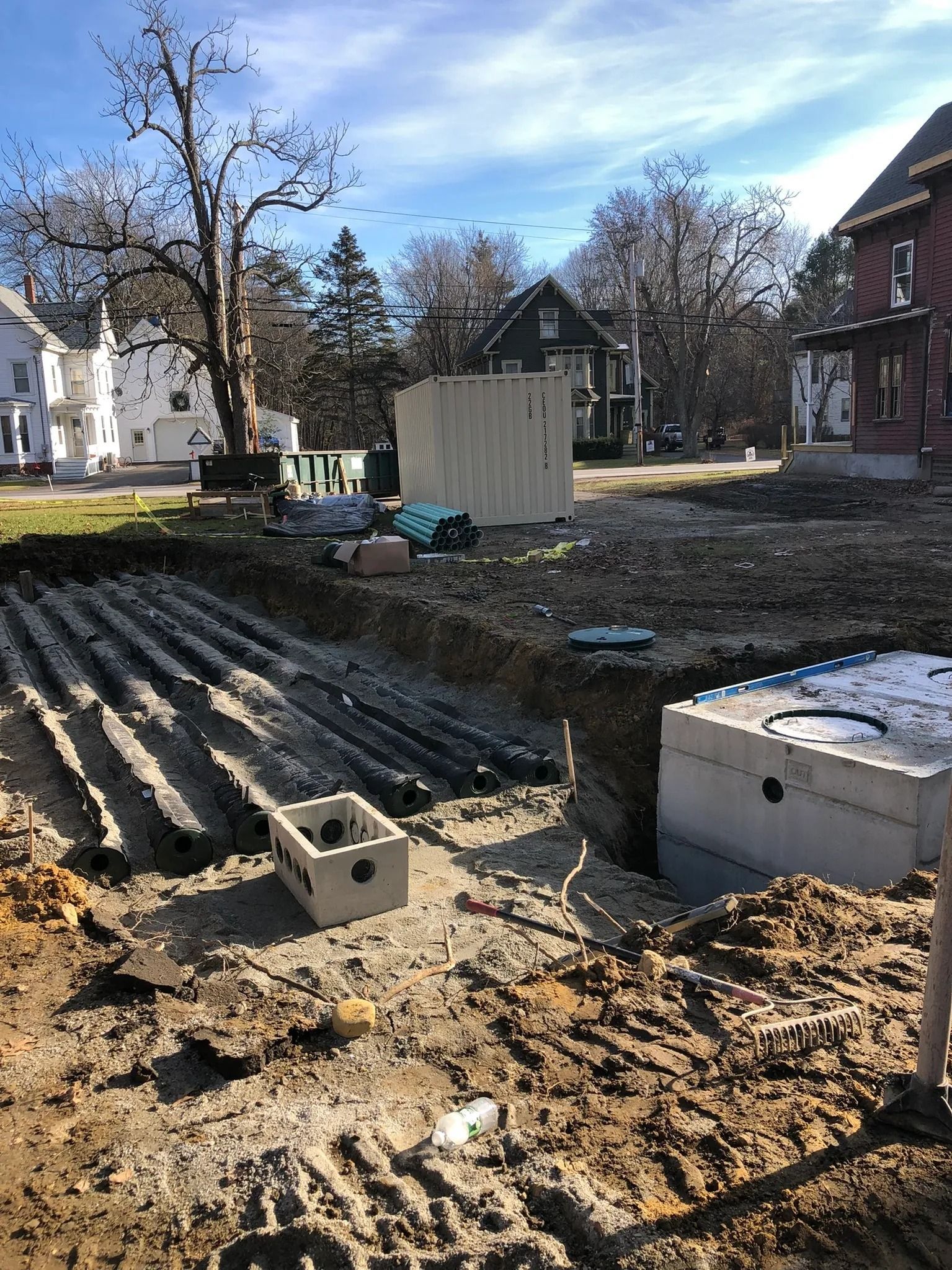Construction site with trenches, pipes, and concrete tanks. Buildings and trees in the background under a blue sky.