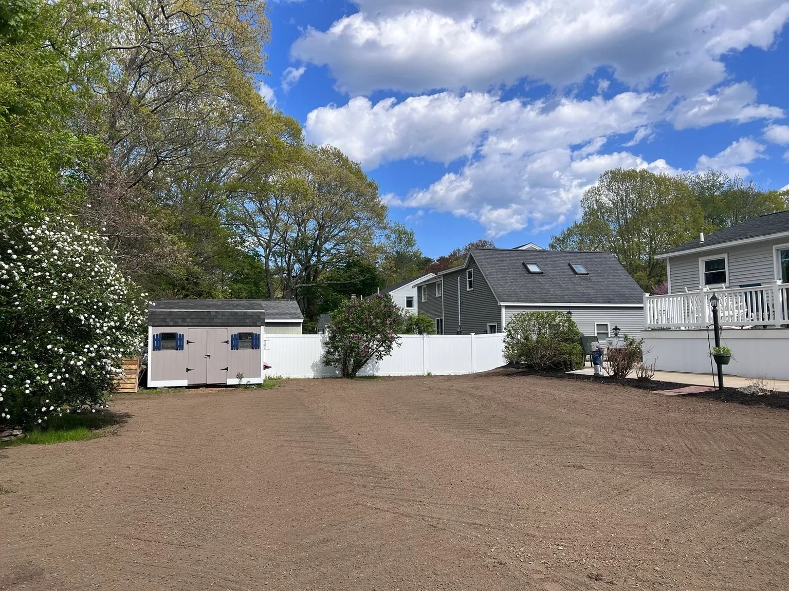 Gravel yard with white fence, shed, and houses under a cloudy blue sky.