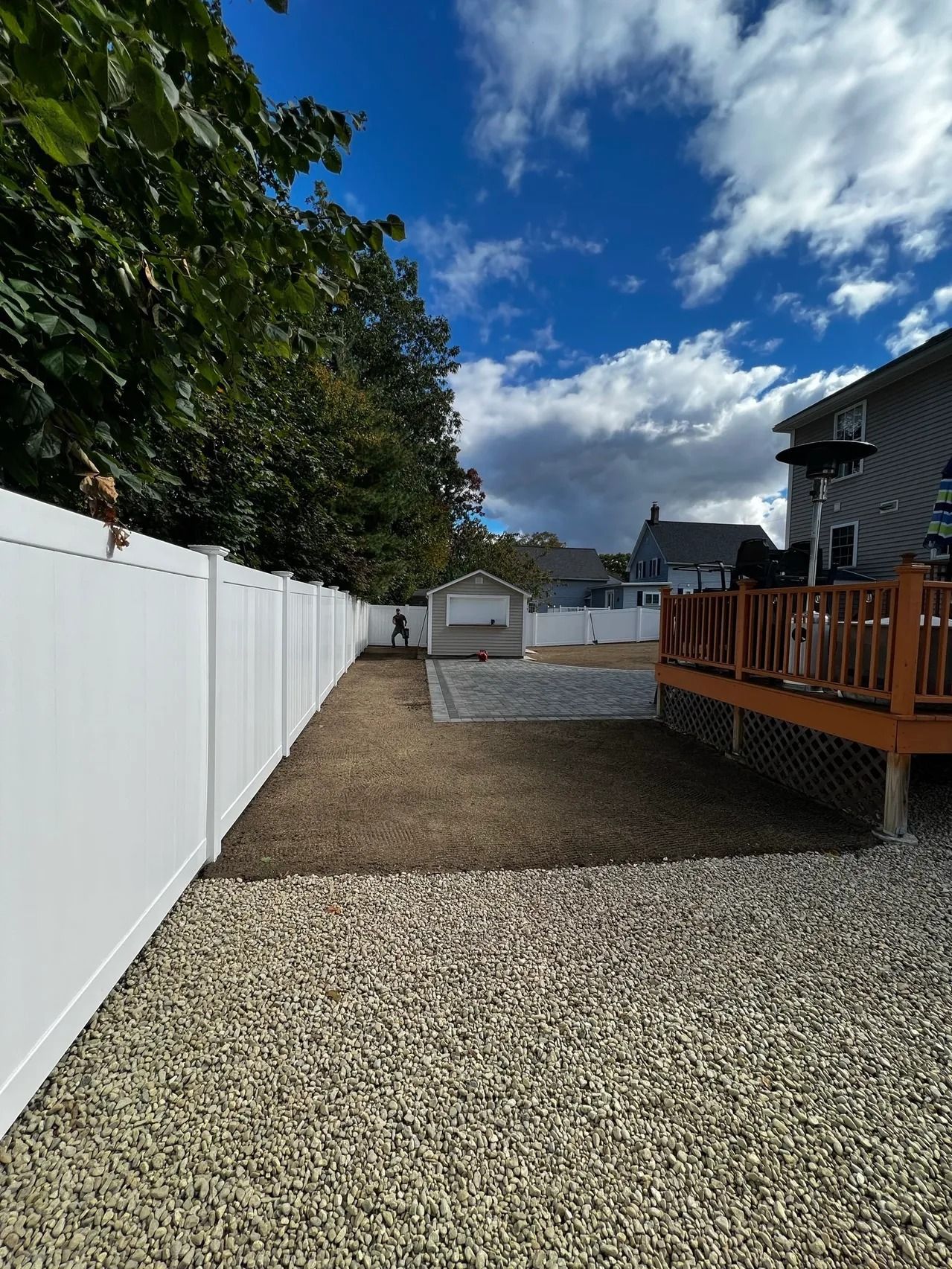 Backyard with white fence, gravel, lawn area, deck, and shed under blue sky.