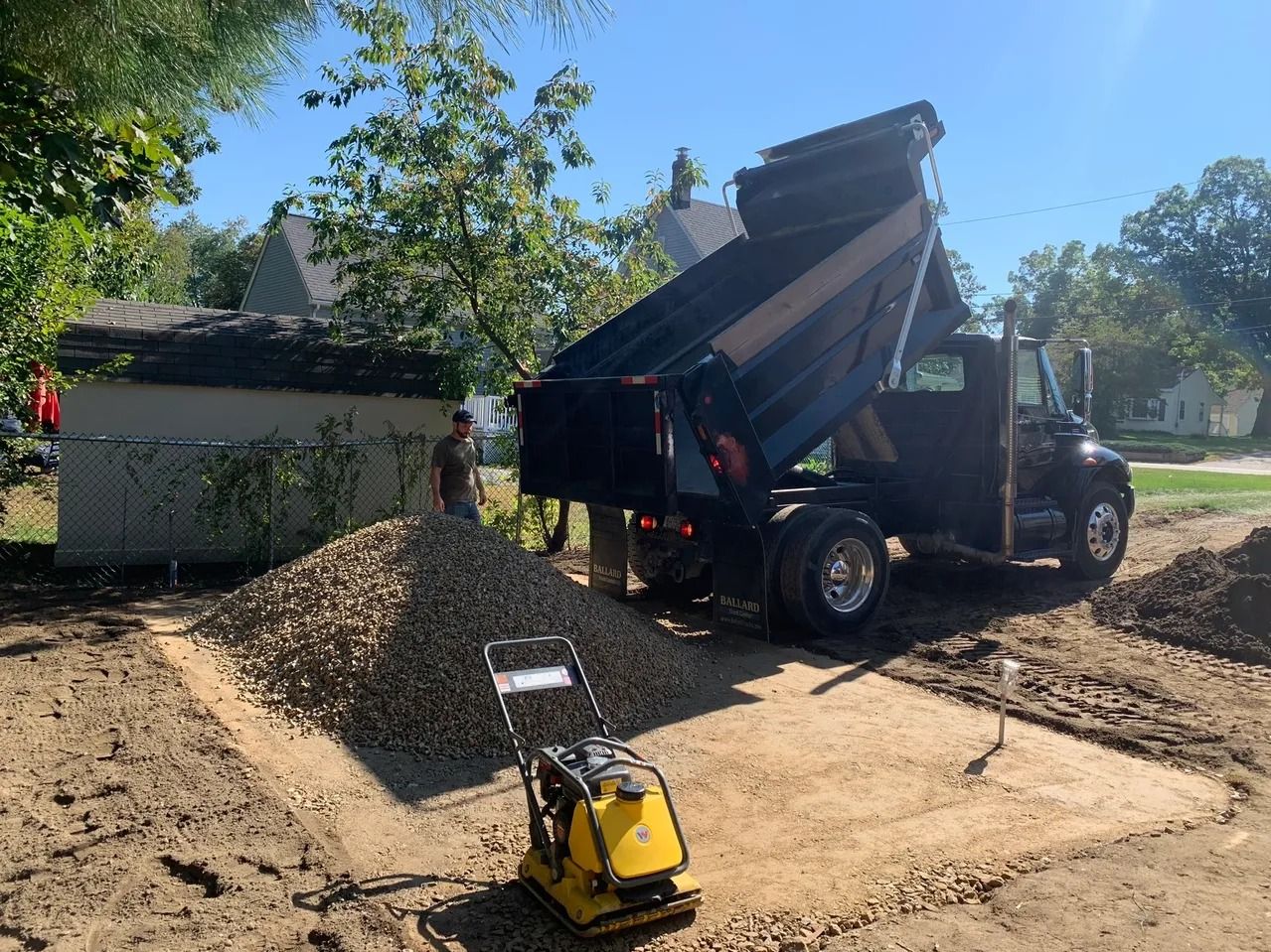 Dump truck dumping gravel onto a prepared ground, a worker nearby. A compactor is in the foreground.