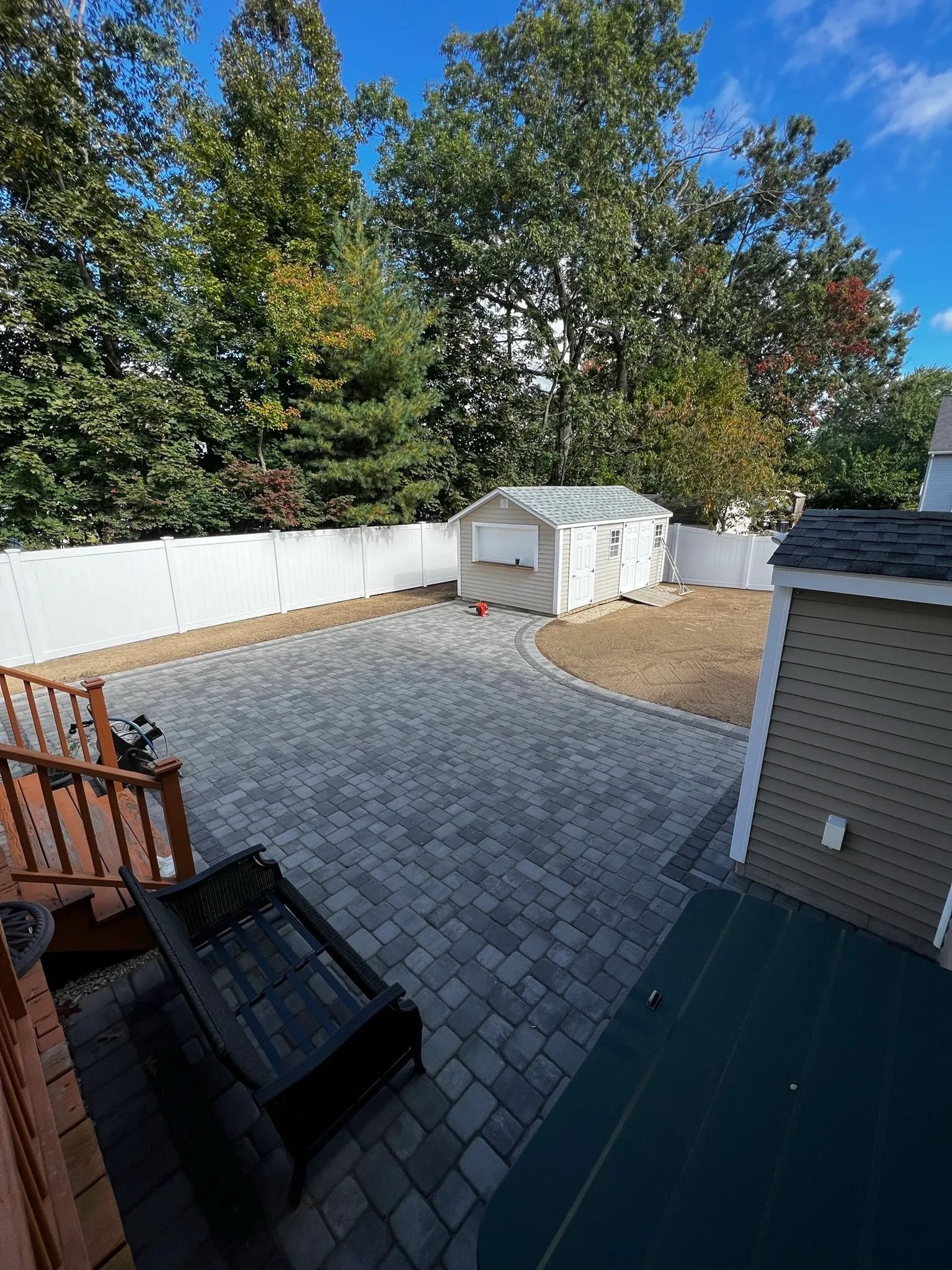 Brick patio with shed in a backyard, surrounded by trees and a white fence.