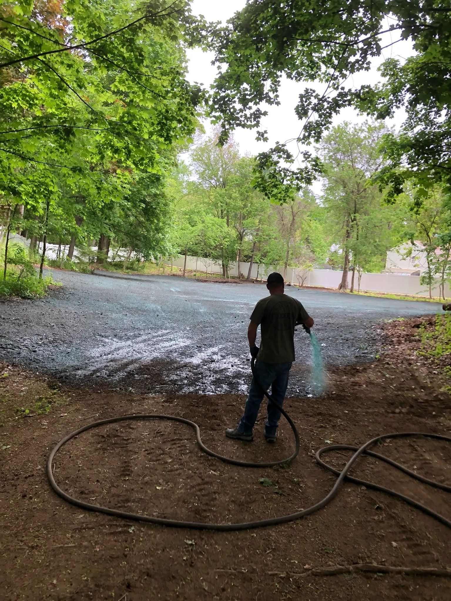 Person spraying water on a gravel lot; trees in the background, overcast sky.
