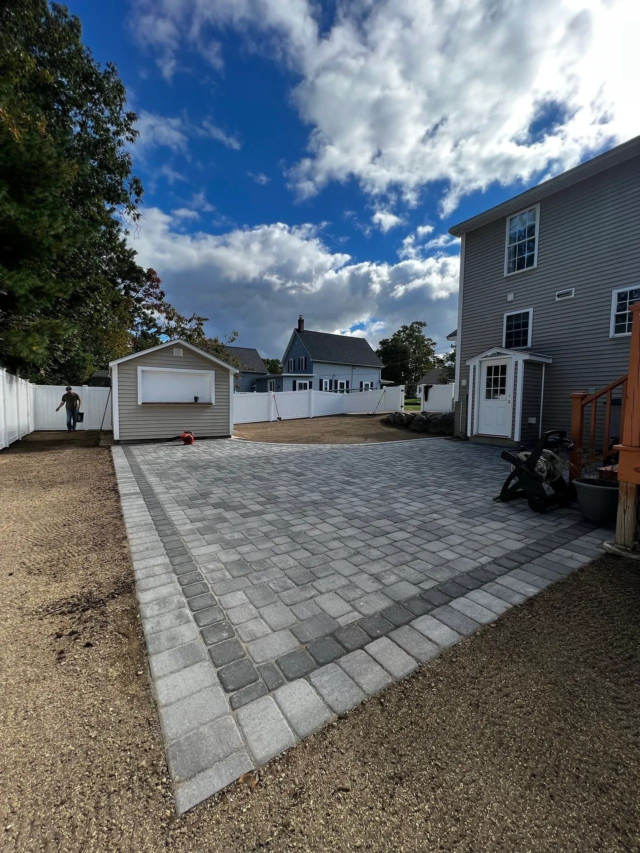 Backyard patio with gray pavers and a bordering of darker pavers, with a white garage and building.
