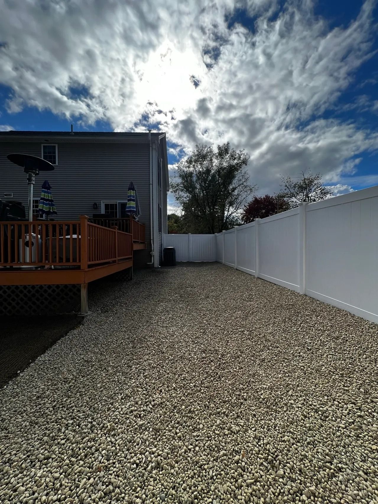 Gravel-covered side yard with white fence and wooden deck next to a house under a cloudy sky.