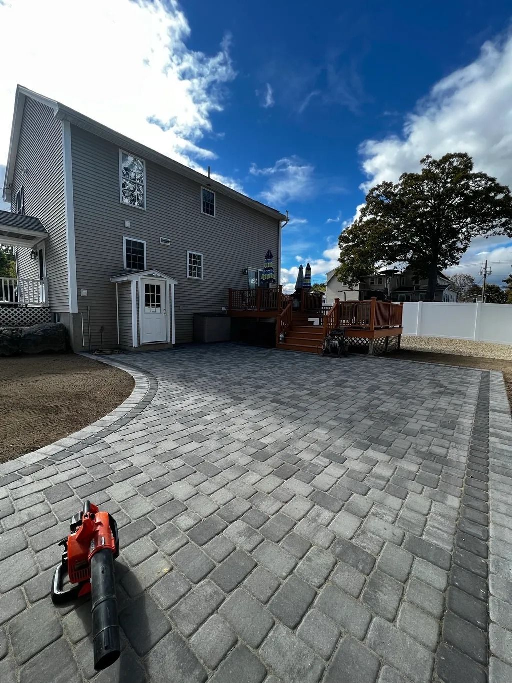 Gray brick paver driveway leads to a house with a wooden deck and a cloudy sky. A leaf blower rests on the pavers.