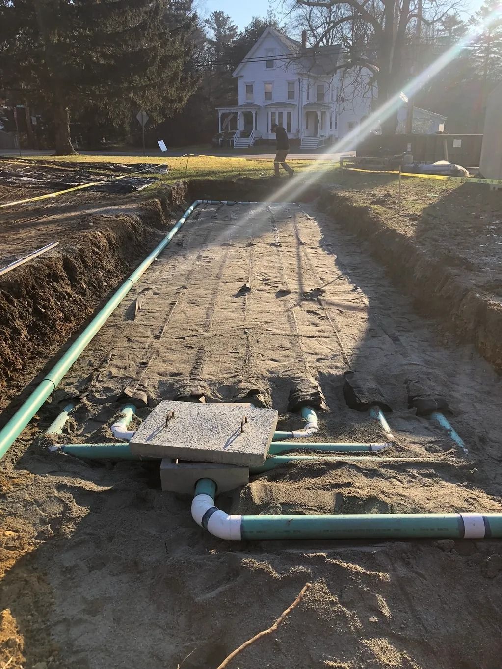Construction of a septic system: green pipes, concrete box, and trenching in a yard with a house in the background.