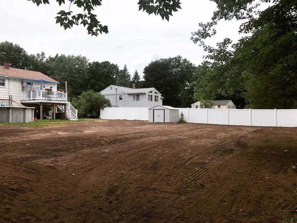 Cleared lot with brown earth, white fence, and houses in the background under a cloudy sky.