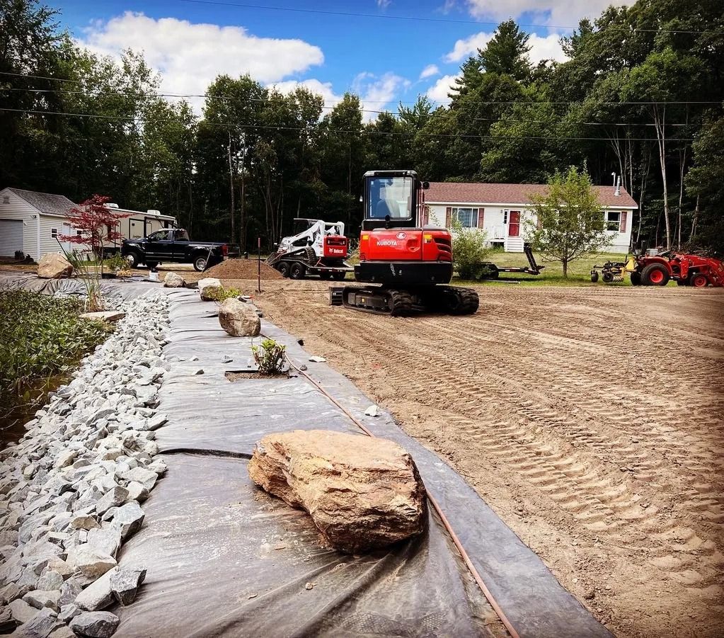 Construction site with excavator and equipment near a house and trees. Ground cover, rock border visible.