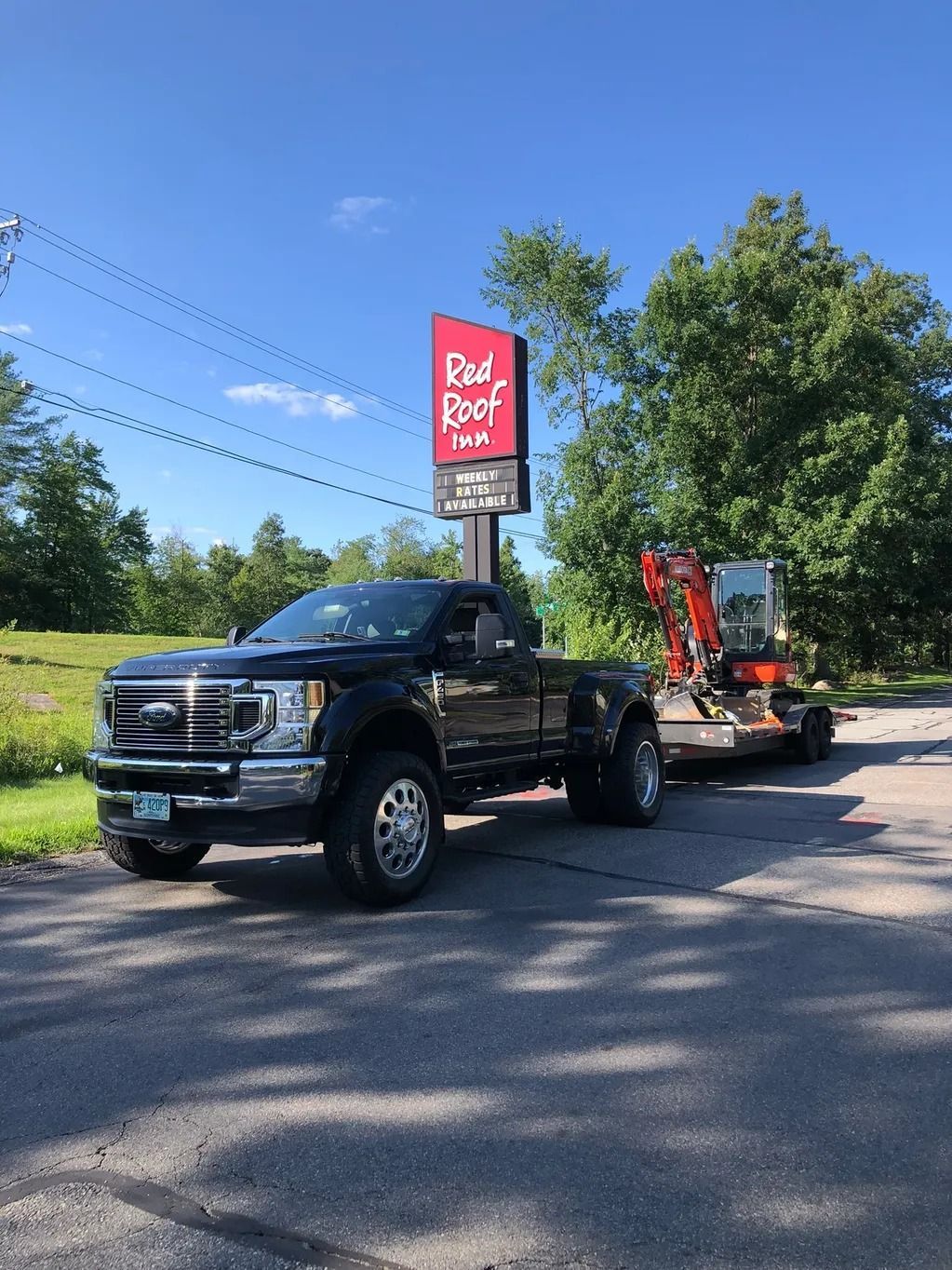 Black truck towing a small excavator on a trailer, parked in front of a Red Roof Inn sign, under a blue sky.