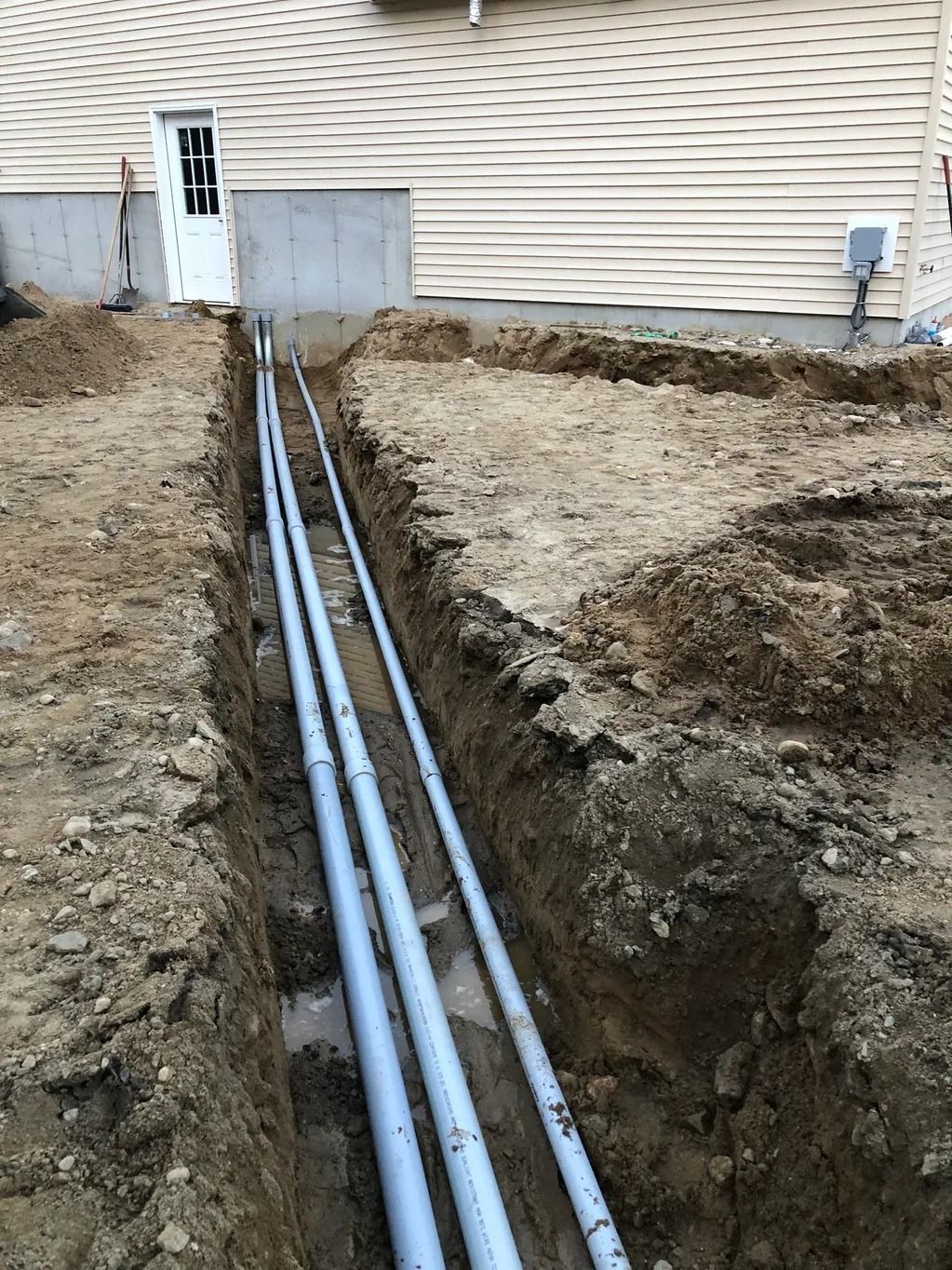 Trench with gray pipes running towards a building, dirt on either side.
