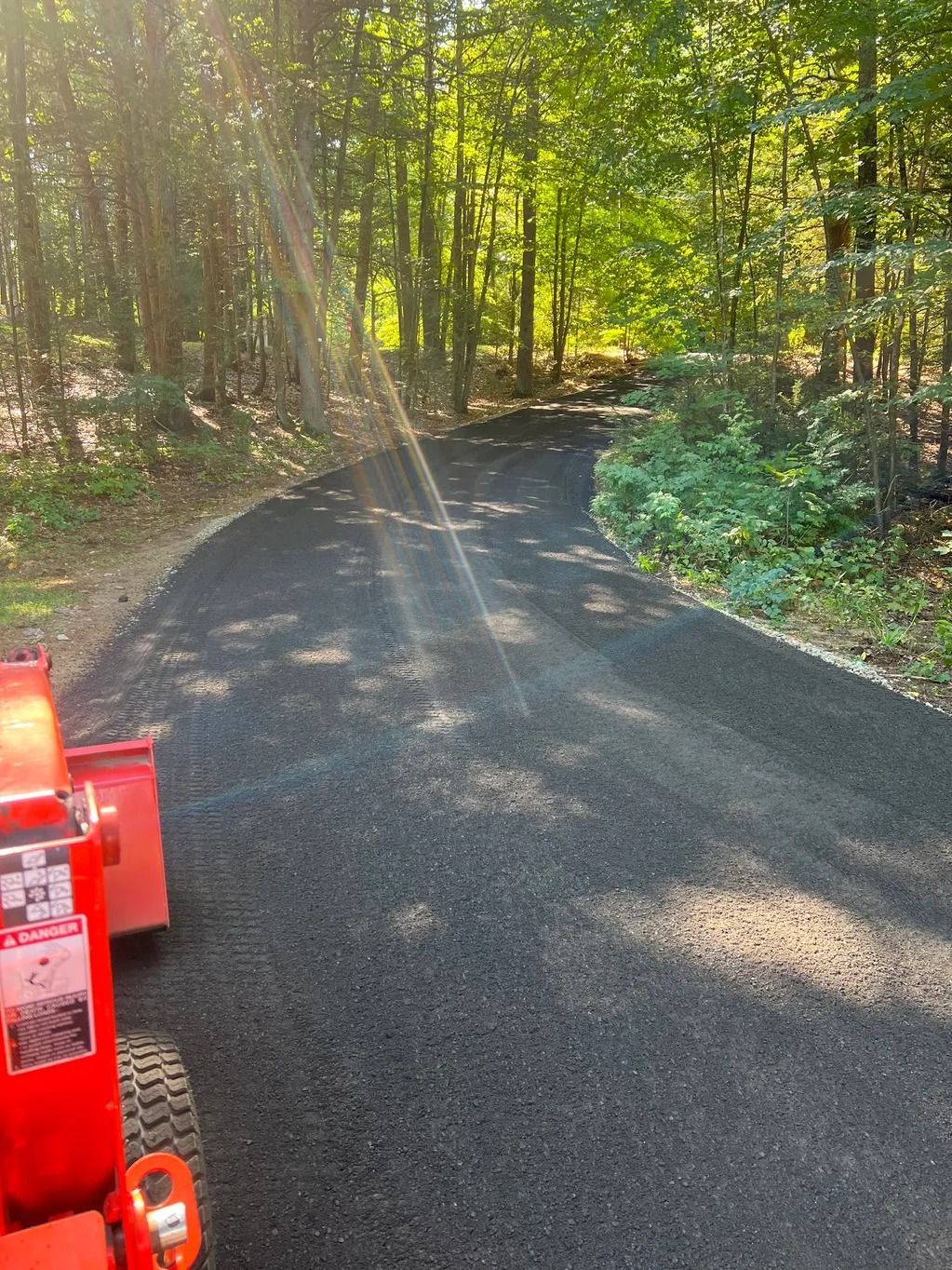 Paved asphalt driveway curves through a wooded area. An orange tractor is visible in the lower left corner.