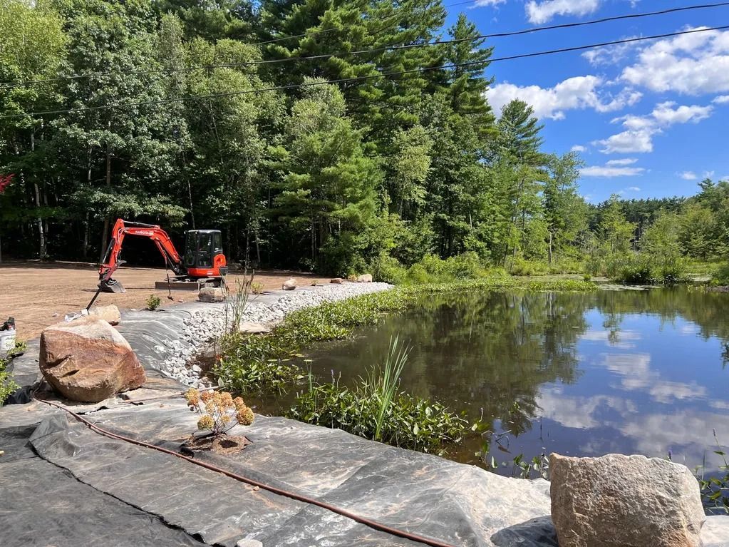 Mini excavator next to a pond. Shoreline reinforced with rocks and cloth. Trees and a blue sky in the background.