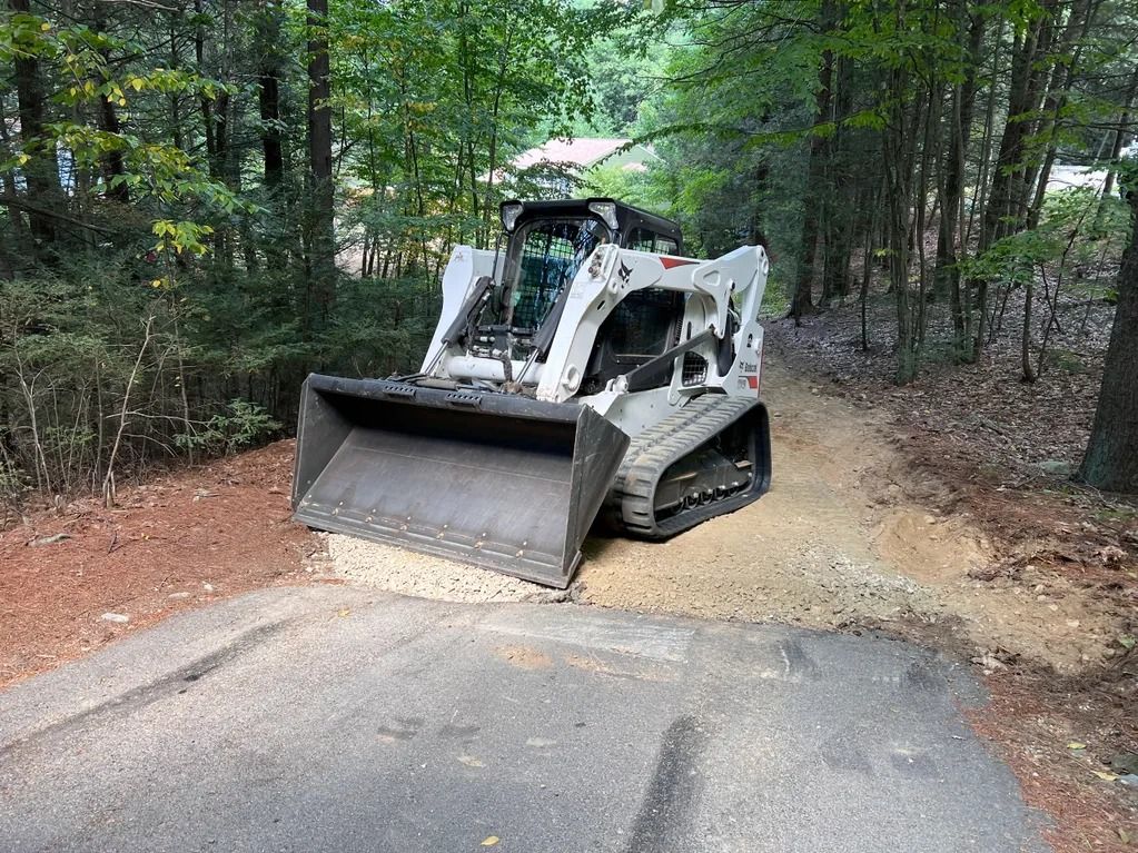 Bobcat skid-steer loader on a path, clearing debris. Trees in the background, a sunny day.