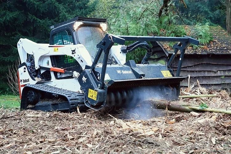 Bobcat skid-steer with a brush cutter mulching wood debris in a forest.