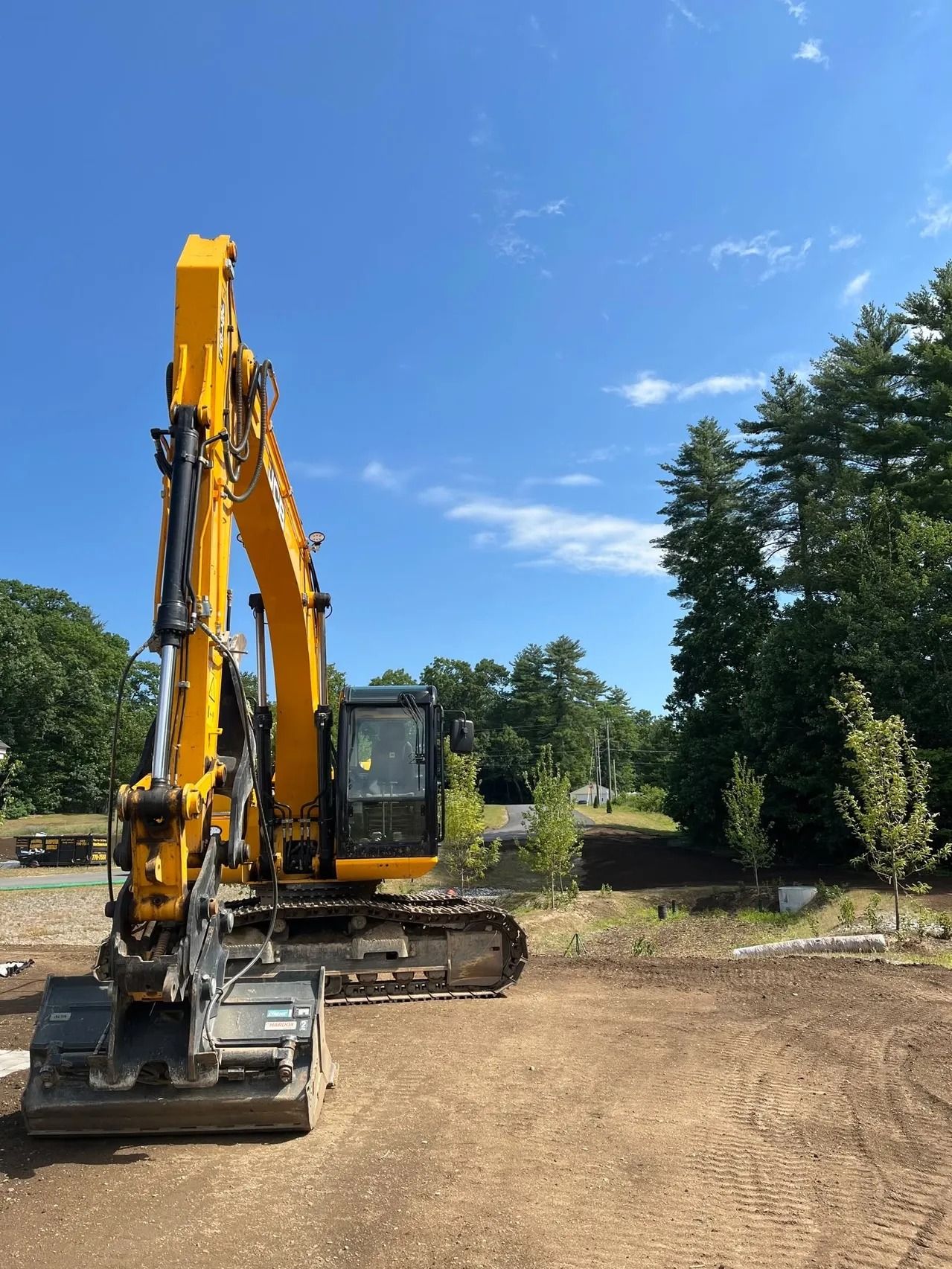 Yellow excavator on dirt, arm raised, under blue sky, near trees.
