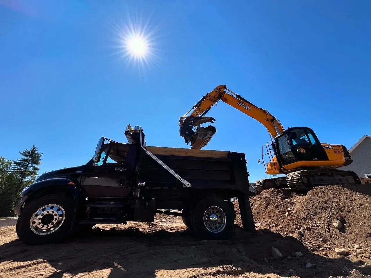 An excavator loading a black dump truck with dirt on a construction site under a bright sun.