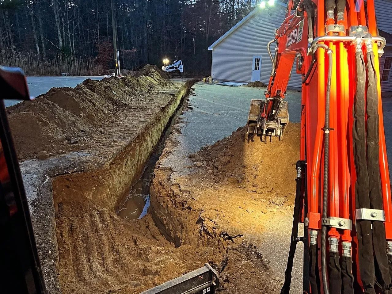 An excavator digs a long trench on a gravel surface near a building at dusk; orange machine, dark soil.