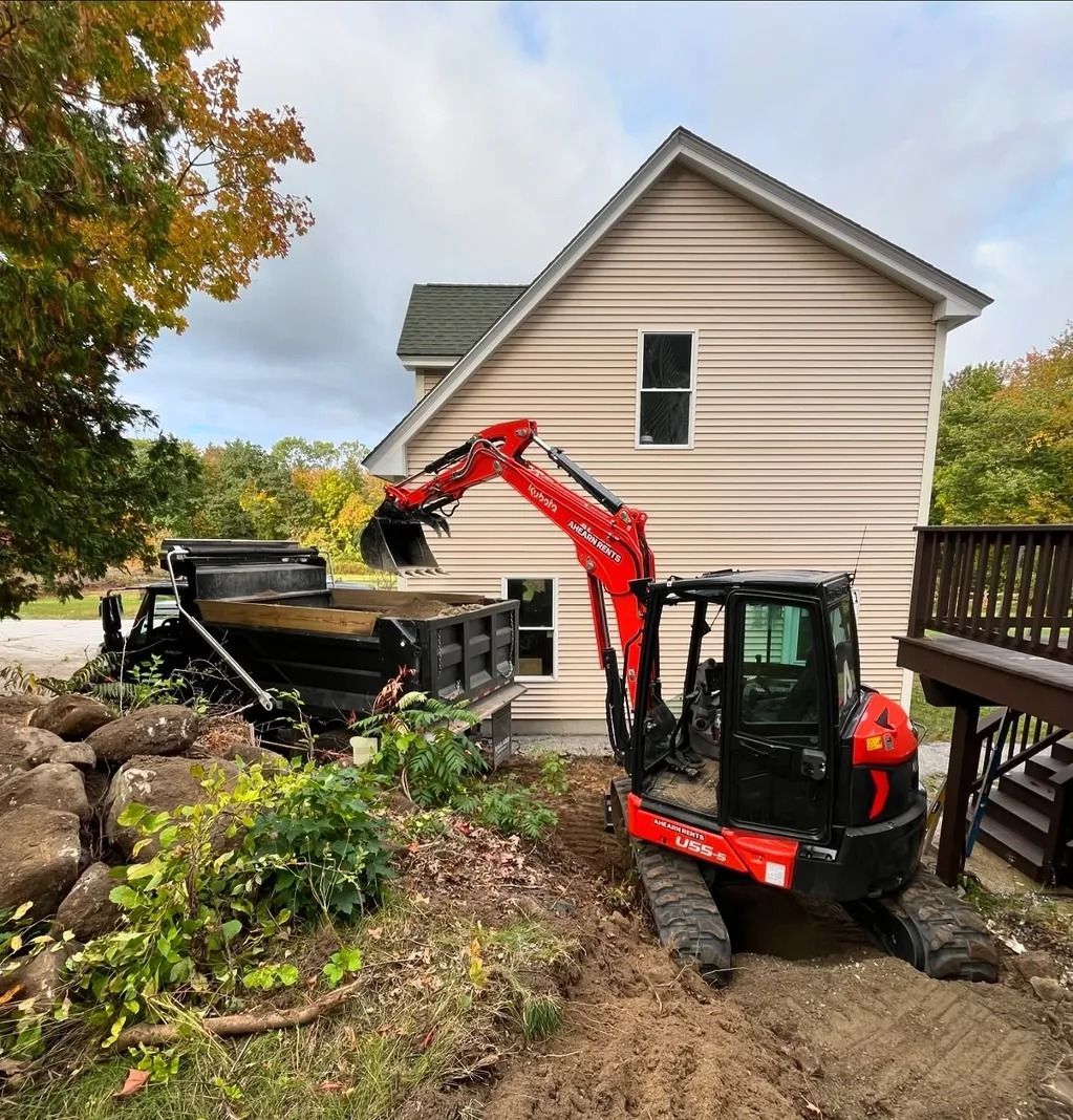 Red excavator loading dirt into a black dump truck next to a beige house, fall foliage in the background.