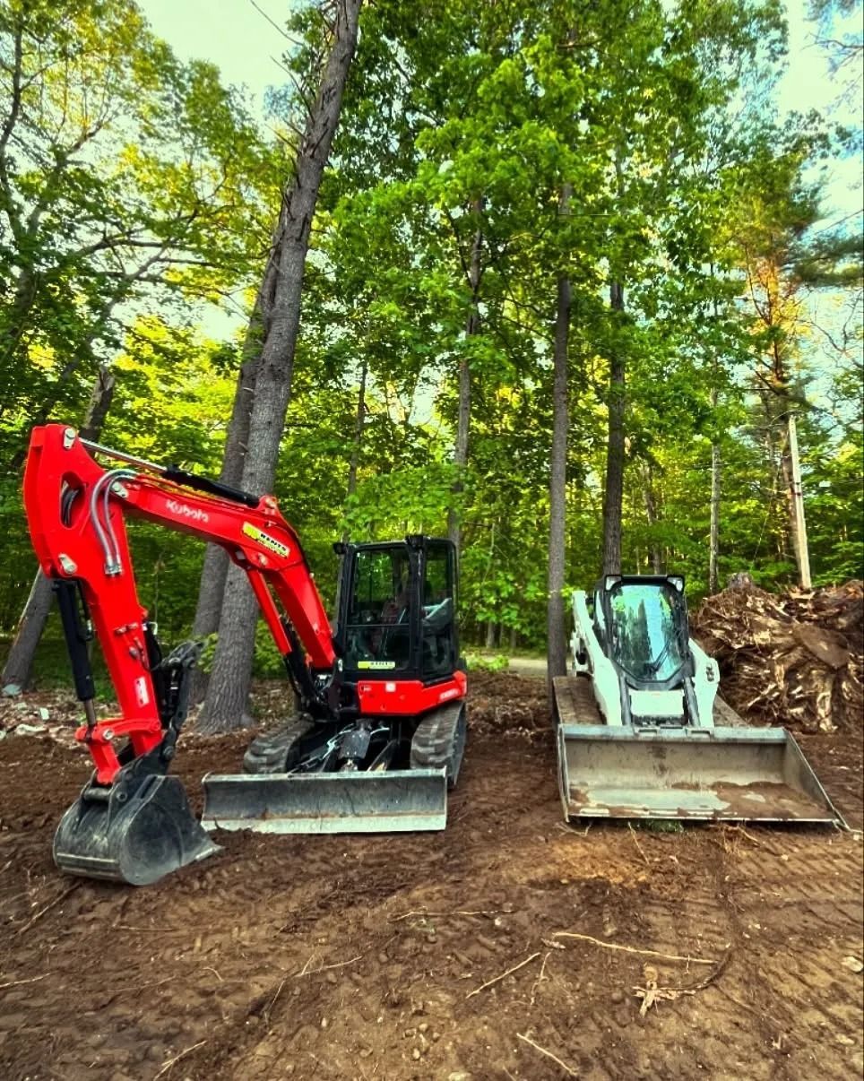 Red excavator and white skid steer on a dirt lot, surrounded by trees.