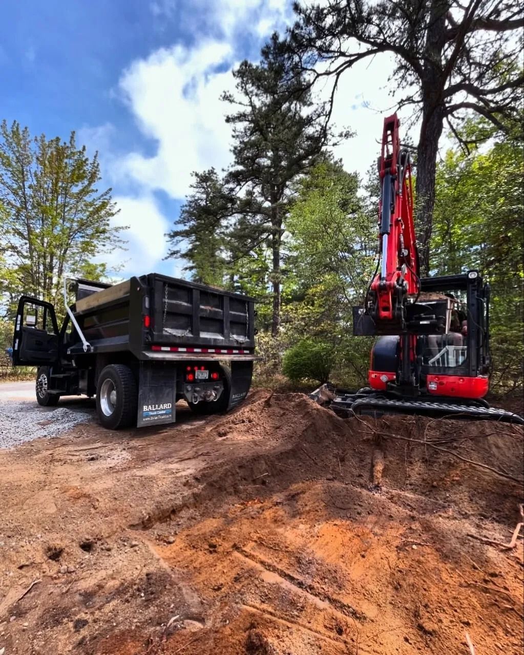 A dump truck and red excavator on a dirt worksite, near trees under a blue sky.