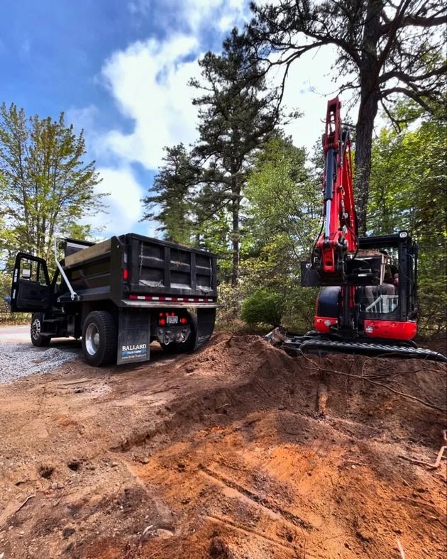 A red excavator digs into dirt beside a black dump truck at a wooded construction site.
