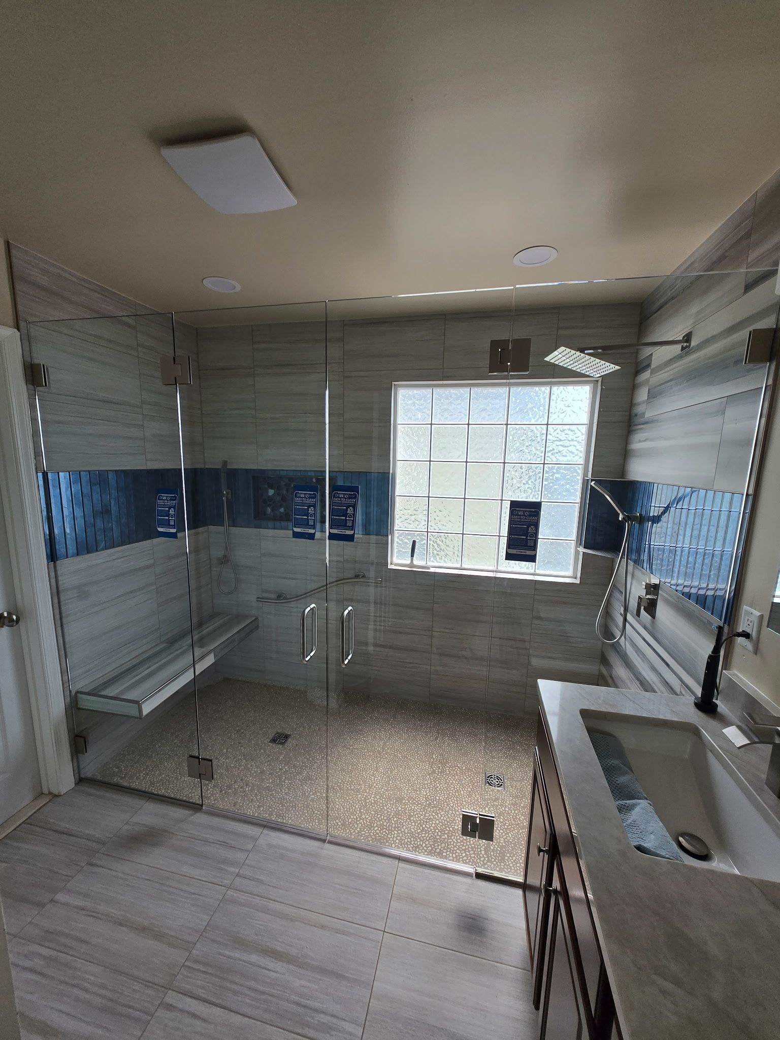 Modern bathroom with glass shower enclosure, blue tile accent, bench, and windowed wall.