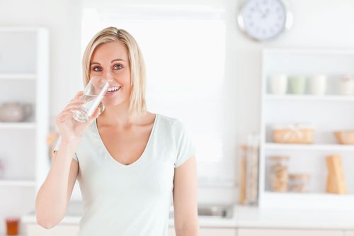 Woman in a bright kitchen drinking from a glass, smiling at the camera.