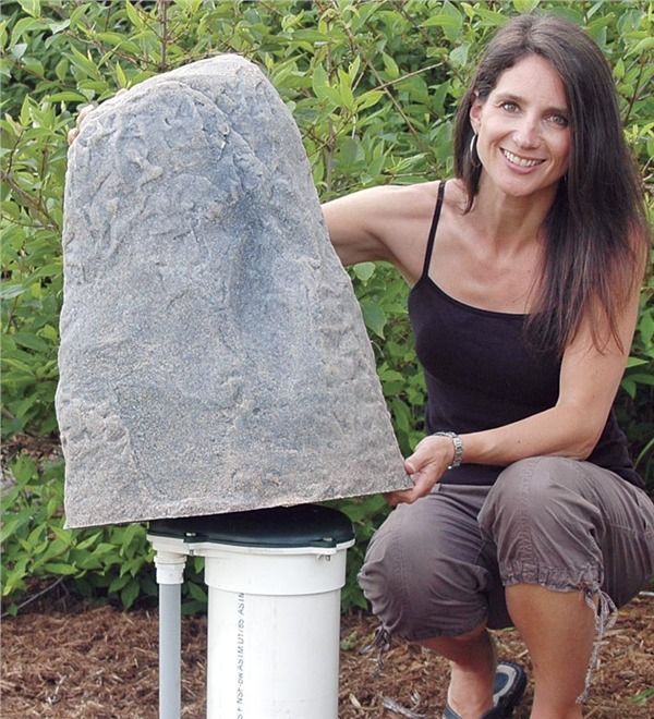 Woman crouches beside a large stone cover on a white outdoor utility post in a garden.