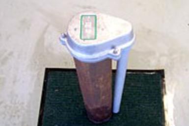 Small trash bin with a white lid, blue handle, and brown body on a dark mat in a corner.