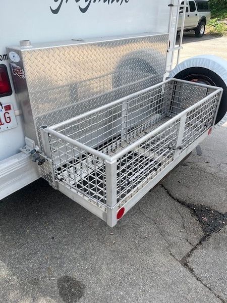 Metal cargo basket attached to the back of a white trailer, next to a silver storage box and spare tire.