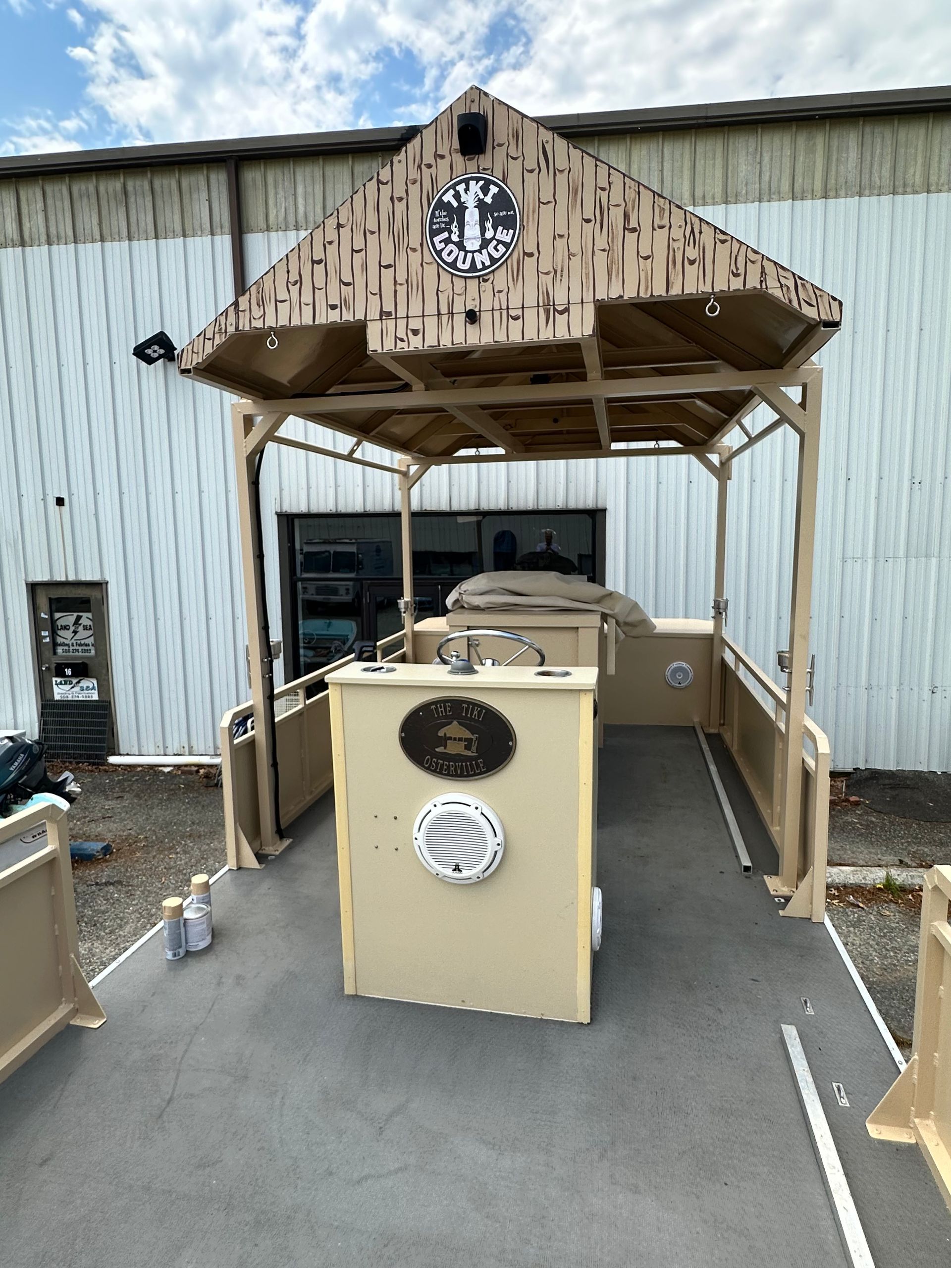 Tan-colored pontoon boat with a covered command center in front of a metal building under a cloudy sky.