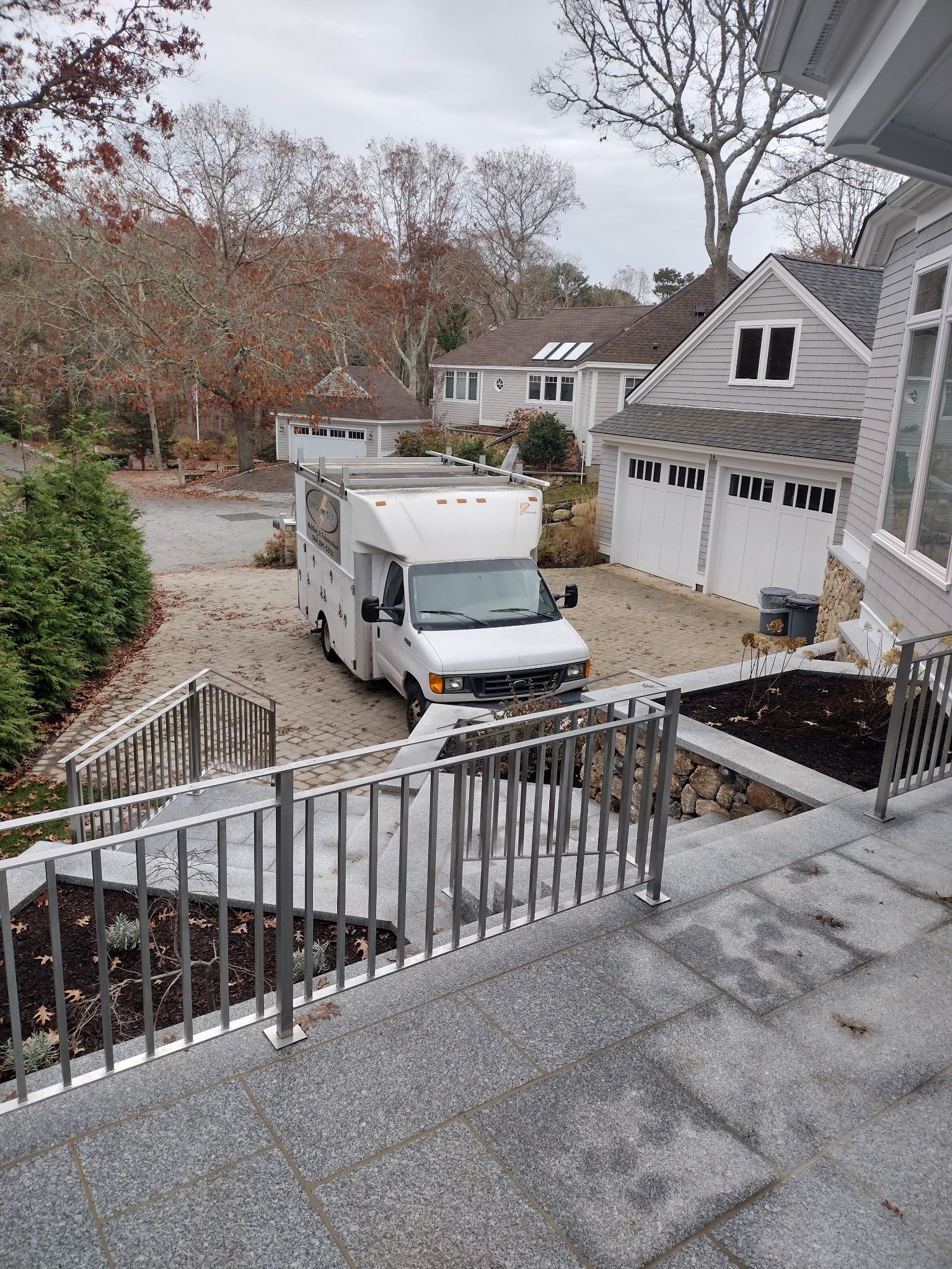 White delivery truck parked in a gravel driveway, steps with metal railings in foreground. Houses and trees in background.