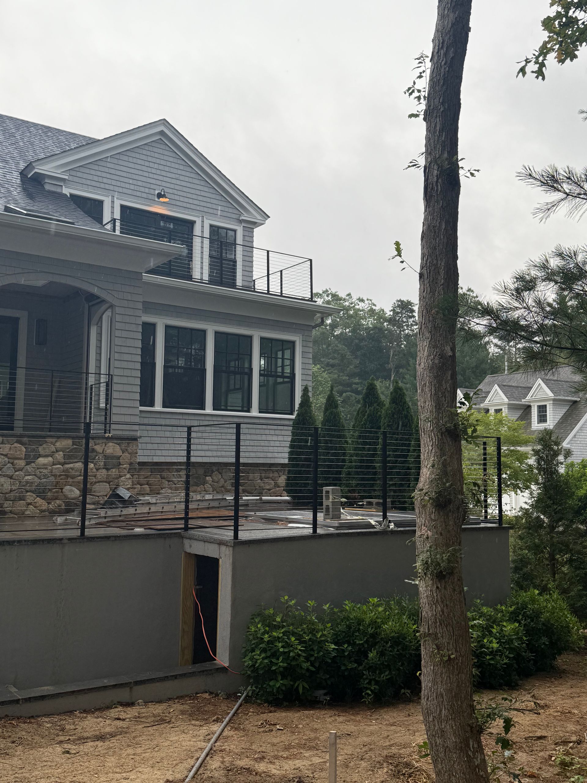 Gray house with stone and cement facade, black railing, balcony, and landscaping under overcast sky.