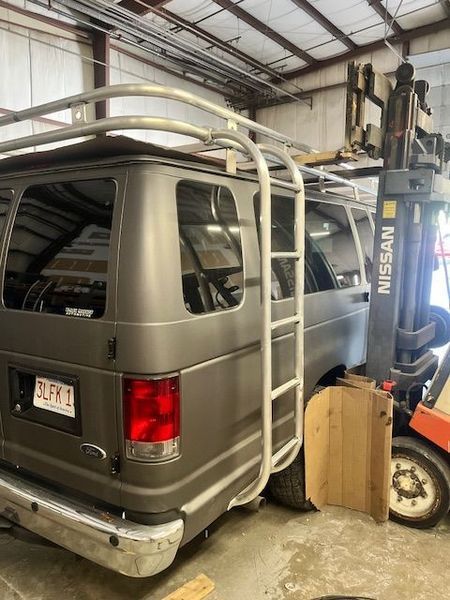 Gray van with ladder and roof rack, parked next to a forklift inside a workshop.