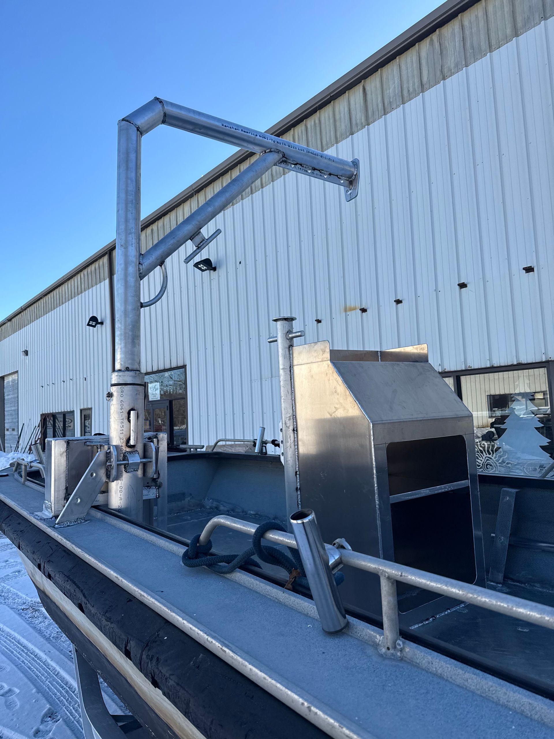 Aluminum boat with a large metallic boom arm attached, parked in front of a corrugated metal building on a snowy day.