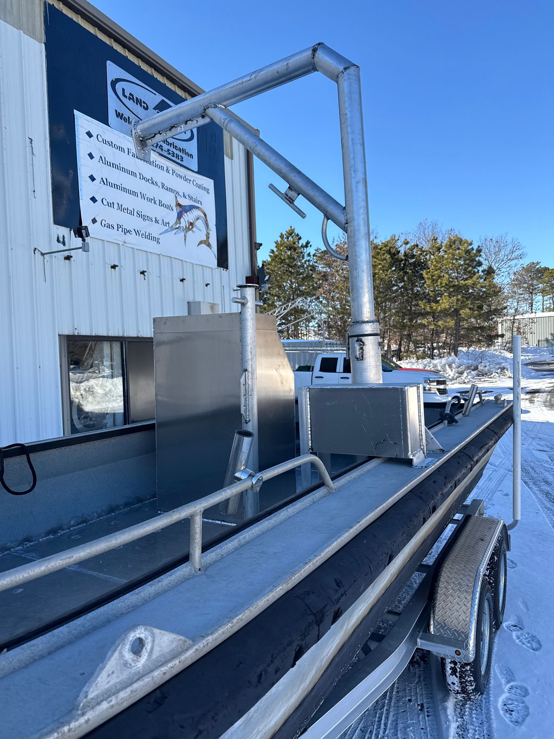 Aluminum fishing boat on a trailer, with a crane attached. Snowy outdoor setting.