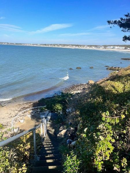 Staircase descends to a rocky beach with ocean view under a blue sky; greenery and a handrail.
