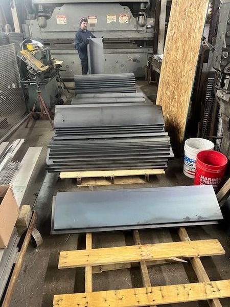 Person in a workshop holding a metal sheet. Steel sheets stacked on pallets. Other tools in background.