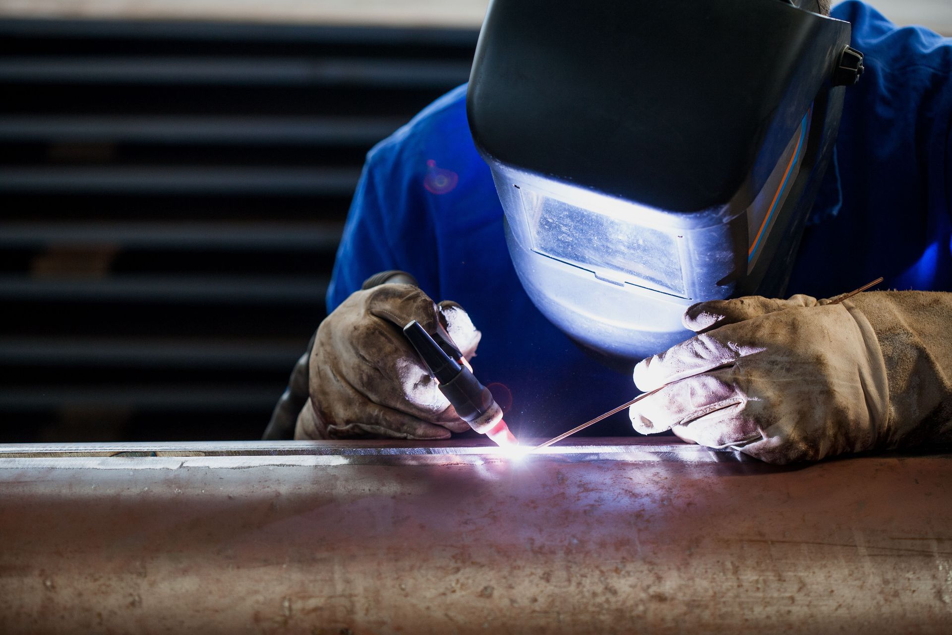Welder in blue work clothes using a welding torch on metal pipe in a workshop.